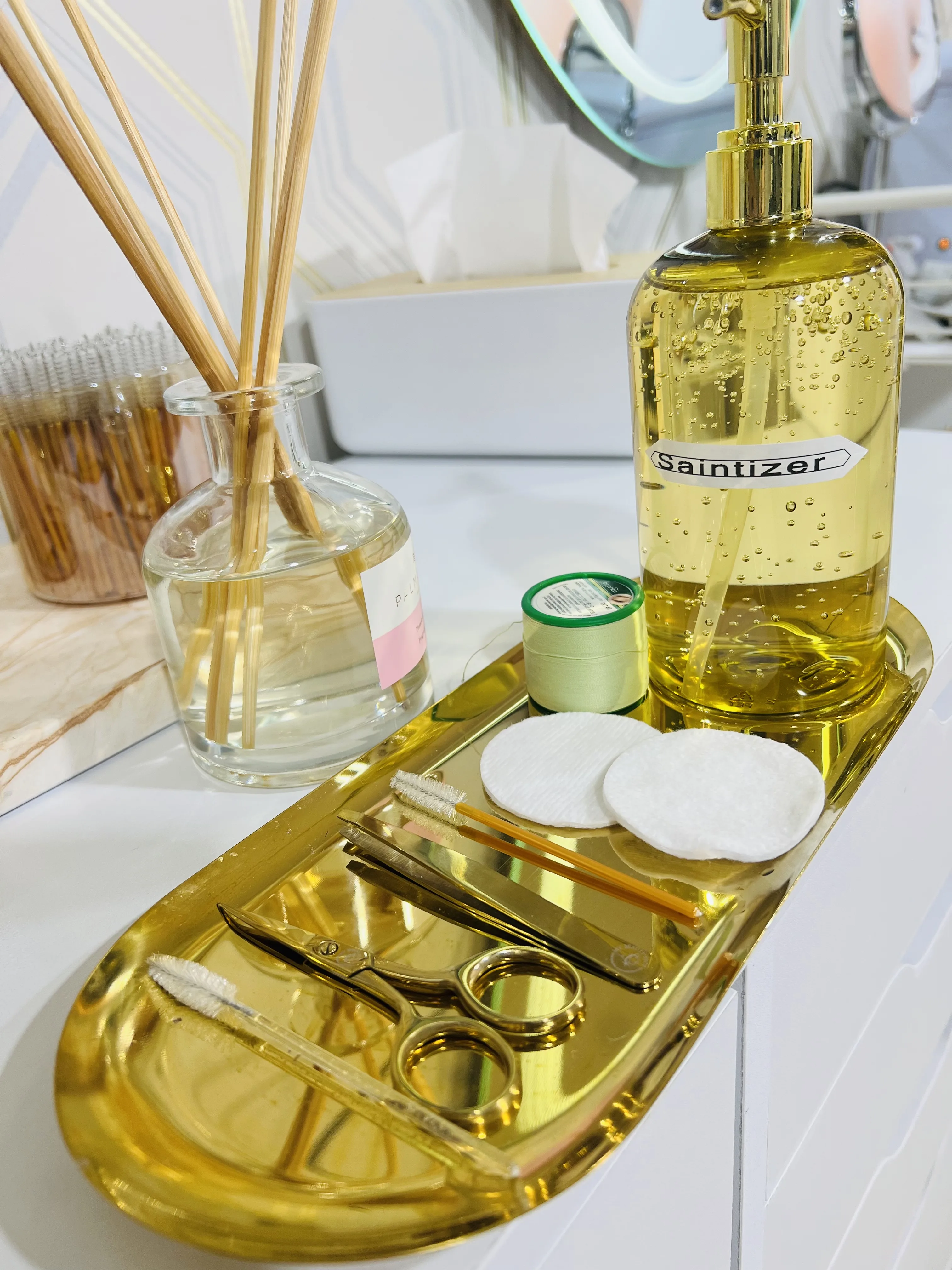 Gold brow tool tray — gold scissors, spoolies, cotton pads, reed diffuser and a sanitiser pump on the preparation station, ring-light mirror behind