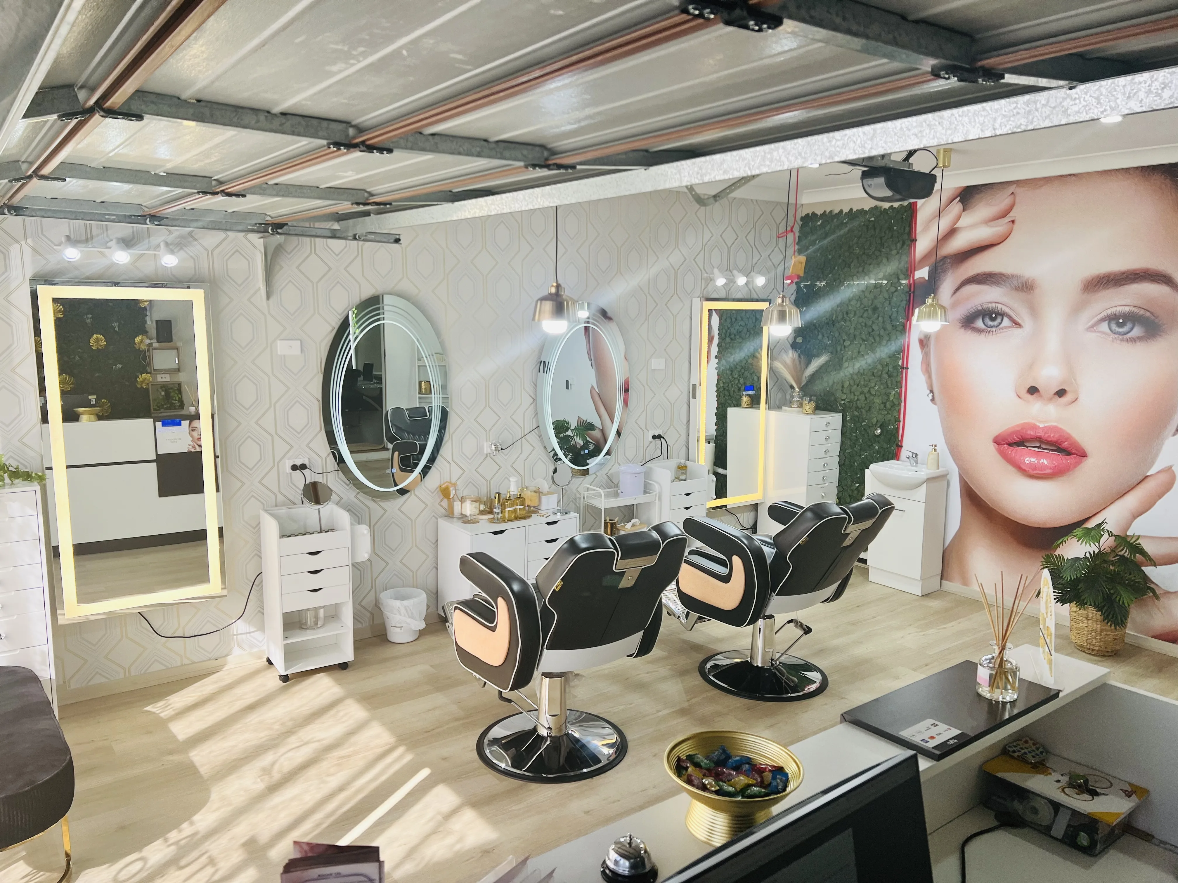 The My Beauty Stop styling floor — two black styling chairs, round backlit mirrors, moss-green feature wall, and gold-topped product shelves