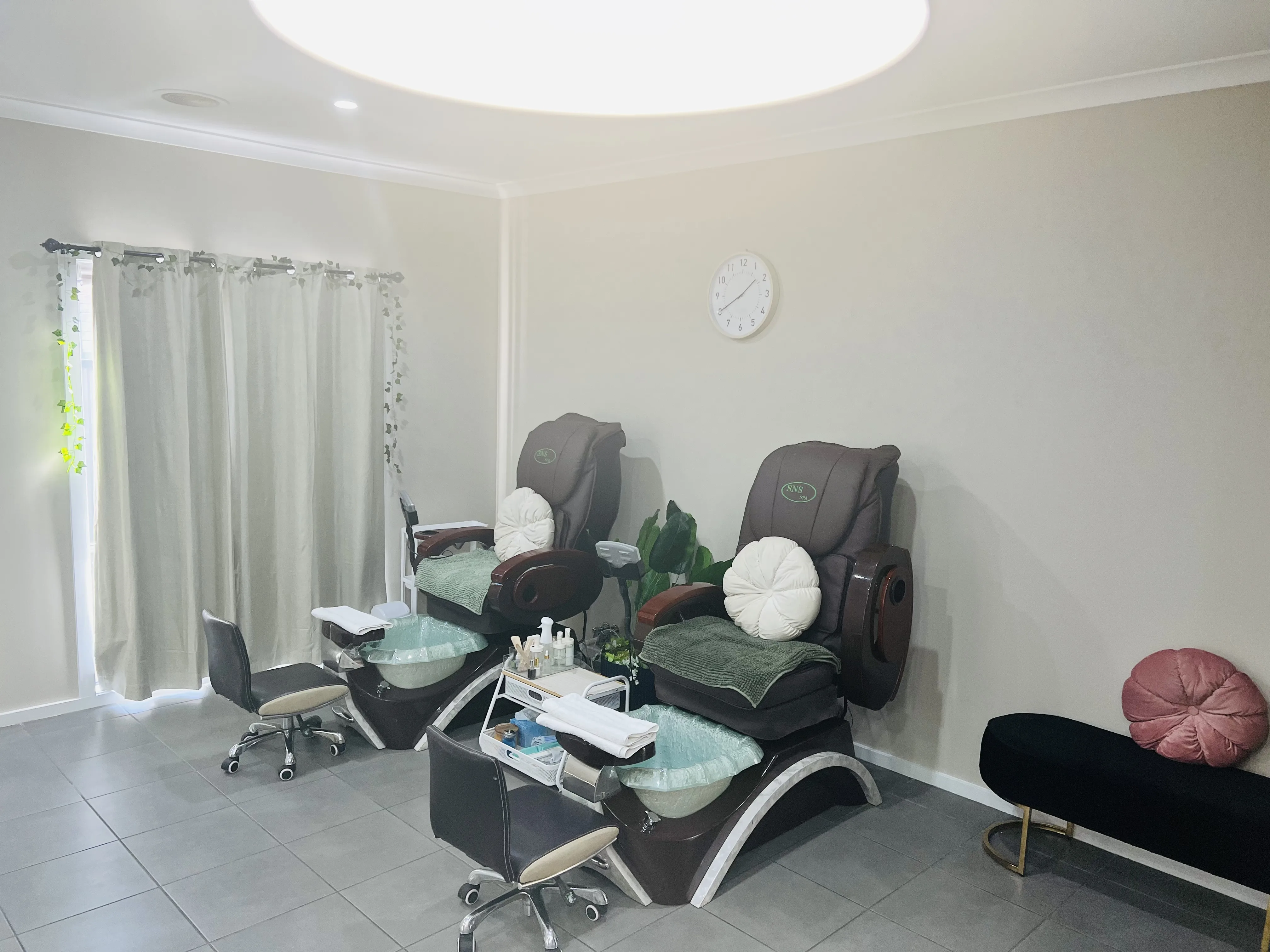The pedicure suite — two SNS spa pedicure chairs with sage towels, sheer curtains, a wall clock, a leafy plant and a round ceiling light-well